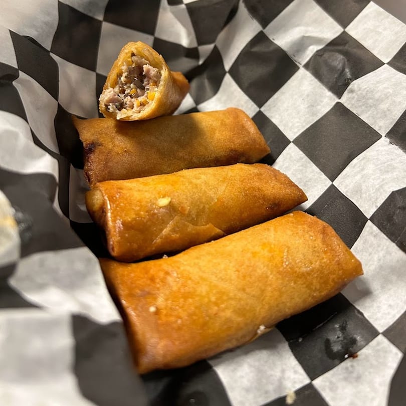 Four golden-brown Vietnamese egg rolls, one broken open to show the savory filling, in checkered paper.