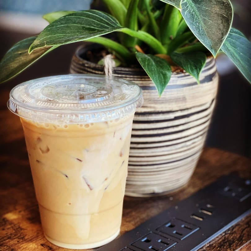 Iced Vietnamese coffee in a plastic cup with a plant and power strip on a wooden table.