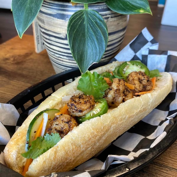 Shrimp banh mi sandwich with cilantro, pickled carrots, cucumbers, and jalapeños in a black basket.